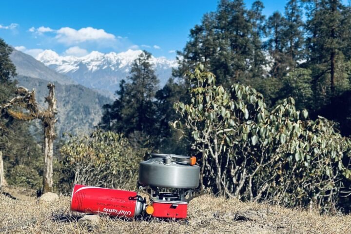 A camping stove and gas canister on dry grass with snow-capped mountains behind