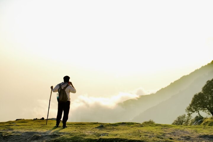 A man carrying a rucksack on mossy ground, leaning on a walking cane while looking at a mountain view