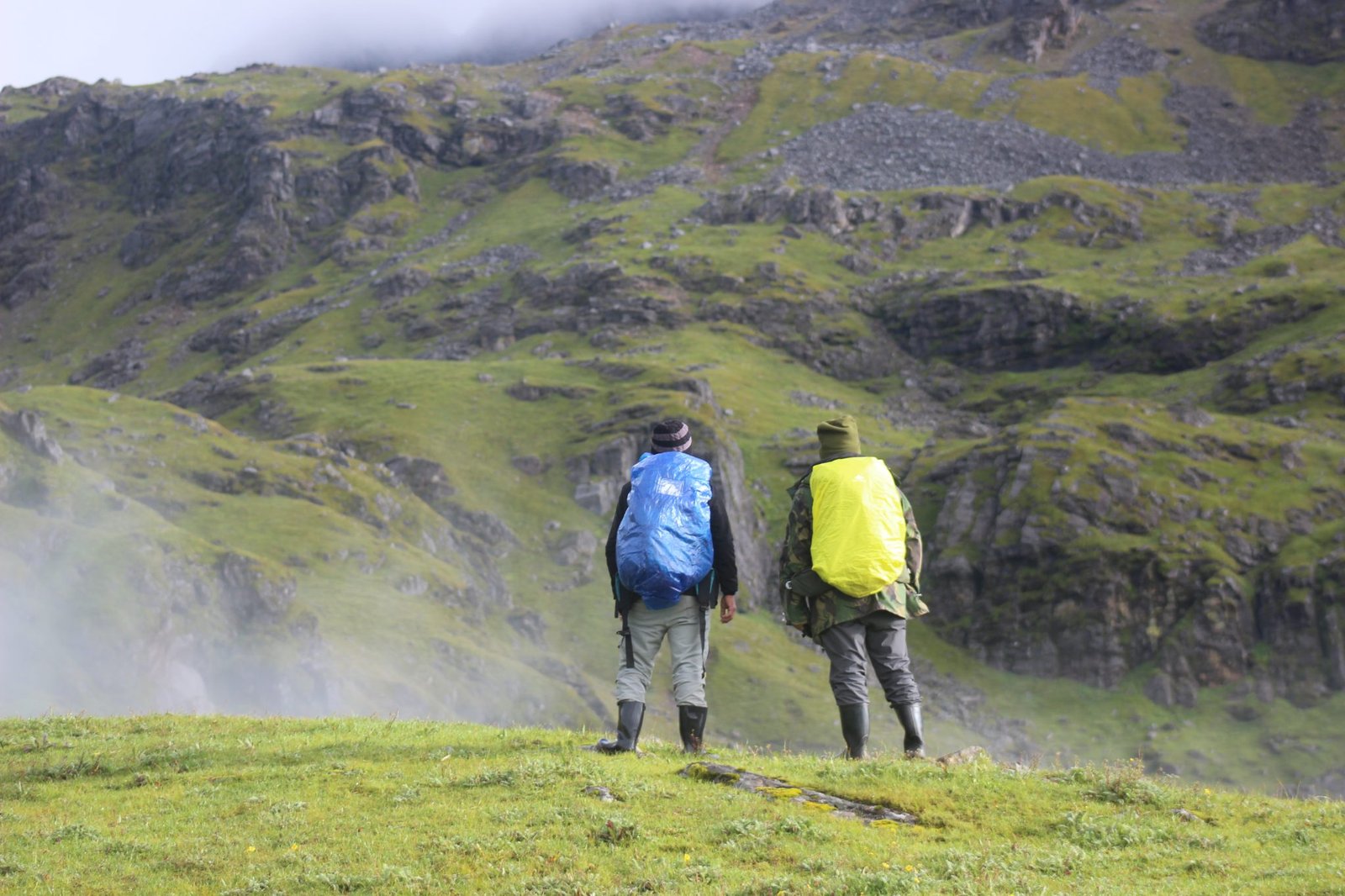 Two hikers wearing backpacks with blue and yellow rain covers on a grassy hillside facing mountains