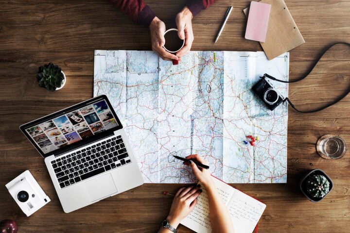 Two people planning a trip at a wooden table with a road map, laptop, camera and notebook