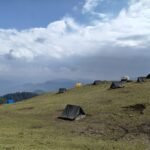 Camping tents pitched on a grassy mountain ridge under dramatic clouds with Himalayan views
