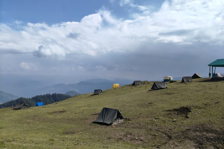 Camping tents pitched on a grassy mountain ridge under dramatic clouds with Himalayan views