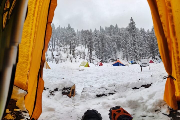 View from inside a tent onto a snowy campsite with tents and snow-covered pine trees