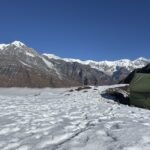 A green camping tent on snow-covered ground below rocky, snow-capped mountains under a clear blue sky