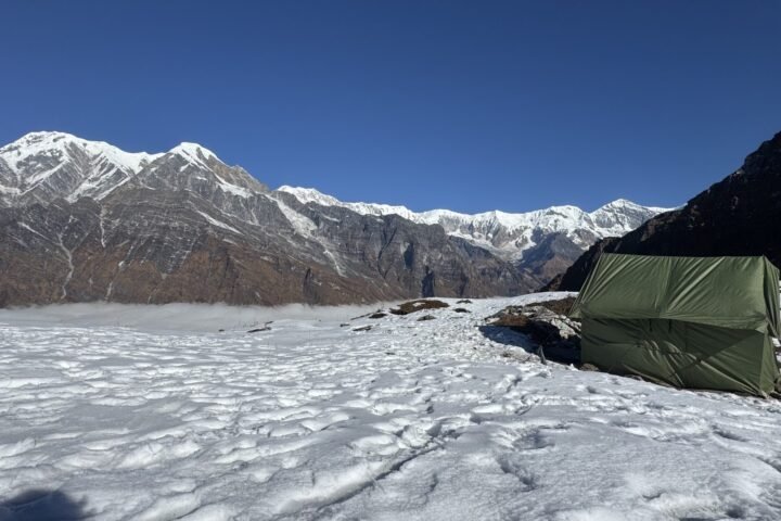 A green camping tent on snow-covered ground below rocky, snow-capped mountains under a clear blue sky