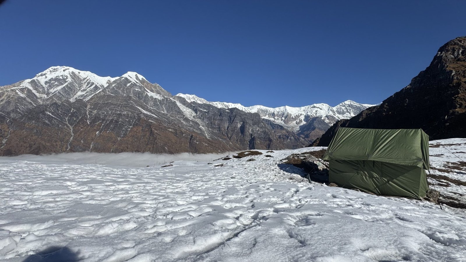 A green camping tent on snow-covered ground below rocky, snow-capped mountains under a clear blue sky